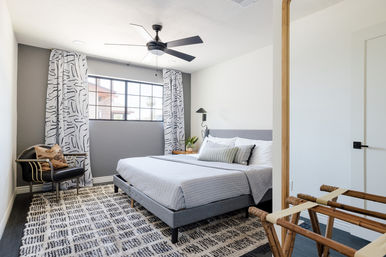 Sunlit modern bedroom with gray upholstered bed and striped bedding, black ceiling fan, large black-framed window with patterned white curtains, leather accent chair, woven luggage rack and textured geometric area rug for a bright, cozy contemporary look.