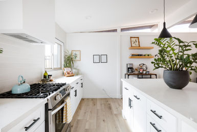 Sunlit modern white kitchen with marble-look island, stainless-steel gas range, light wood floors, large potted plant and open shelves