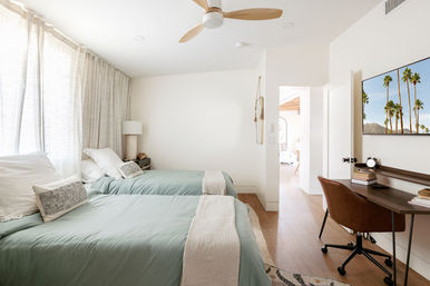 Bright, light-filled modern bedroom with two twin beds in seafoam-green bedding, sheer curtains, wooden ceiling fan, small nightstand, leather desk chair at a workspace and a wall-mounted TV showing palm trees.