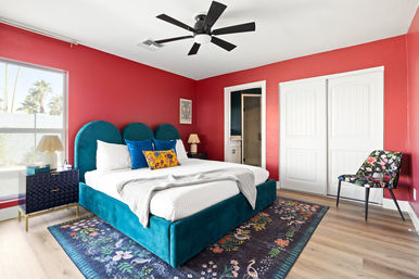 Sunlit modern bedroom with teal velvet bed and rounded headboard, white linens and colorful throw pillows, red accent walls, black ceiling fan, floral area rug, and a window showing palm trees outside.