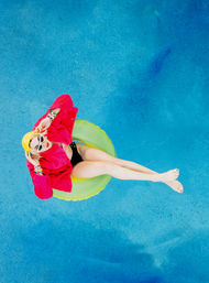 Top-down view of a person in white sunglasses and a hot-pink jacket lounging on a lime-green pool float in a bright blue swimming pool, legs outstretched for a playful summer vibe.