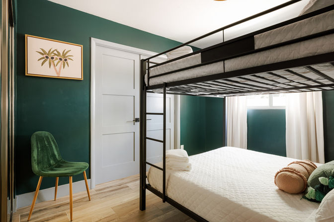 Cozy modern bedroom with black metal bunk bed and white quilted lower mattress, deep green walls, green velvet chair, light wood floors, framed palm art and sunlit window.