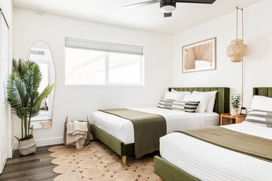 Bright airy twin bedroom with green velvet headboards, white linens and olive throws, striped accent pillows, woven rug, potted plant and full-length mirror.