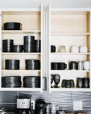 Modern home kitchen with open white cabinets neatly holding stacked matte black plates and bowls, glassware and mugs above a coffee station on the countertop.