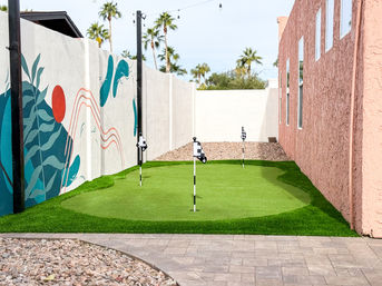 Backyard artificial putting green with three checkered flags between a pink stucco house and a colorful mural wall, palm trees and desert landscaping in the background.