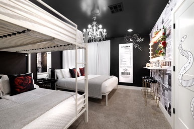 Chic boutique-style guestroom with white bunk bed and queen bed, black-and-white decor, crystal chandelier, lighted vanity with roses, newspaper-print accent wall, red rose throw pillows and gray carpet.
