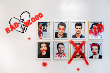 Two rows of four framed portrait photos on a white gallery-style wall defaced with red spray paint and lipstick: dripping paint over eyes and mouths, a large red X over one frame, “SUX” scrawled on another, smeared lipstick marks and red splatters, plus a broken-heart doodle labeled “BAD BLOOD.”