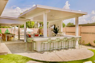 Sunny backyard covered outdoor bar and patio with a long white counter, six mint-green bar stools, built-in grill, lounge seating, dining table and pool umbrella.