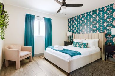 Sunlit modern bedroom with teal bird-patterned accent wall, beige upholstered bed dressed in white linens with a teal throw and decorative pillow, matching teal curtains, black nightstands with lamps, blush armchair, hanging plant, ceiling fan, and light wood floors.