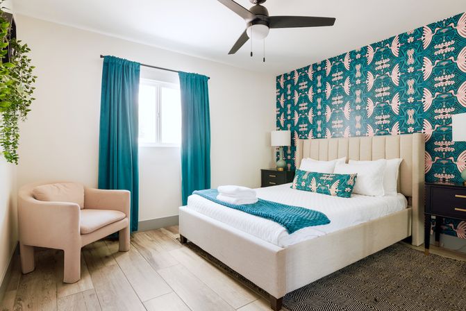 Sunlit modern bedroom with teal bird-patterned accent wall, beige upholstered bed dressed in white linens with a teal throw and decorative pillow, matching teal curtains, black nightstands with lamps, blush armchair, hanging plant, ceiling fan, and light wood floors.