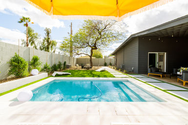 Sunny suburban backyard with a rectangular turquoise pool, yellow umbrella fringe overhead, artificial-turf lawn, lounge chairs, modern dark-gray patio, lone shade tree and cinderblock garden wall.