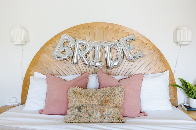 Bridal suite bed with a carved wooden sunburst headboard, silver foil balloons spelling BRIDE, white bedding, pink and textured throw pillows, wall sconces and a potted fern — modern wedding bedroom decor
