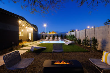 Cozy modern backyard pool and patio at dusk — illuminated rectangular pool, lounge chairs under a yellow umbrella, pergola lights, artificial turf, and a glowing fire pit.