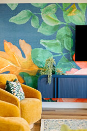 Vibrant living room with oversized watercolor botanical mural, mustard velvet armchair with floral cushion, navy ribbed floating console topped with greenery and decor, hardwood floor and patterned rug.