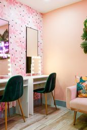 Bright pink makeup vanity with flamingo wallpaper, two lighted mirrors, emerald green velvet chairs, wood floors and a pastel armchair with a tropical-print cushion