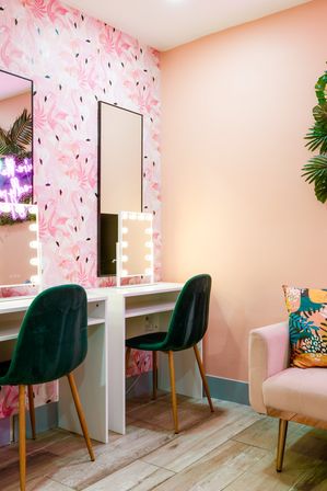 Bright pink makeup vanity with flamingo wallpaper, two lighted mirrors, emerald green velvet chairs, wood floors and a pastel armchair with a tropical-print cushion
