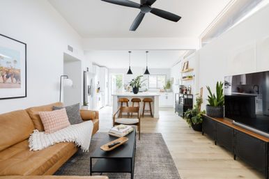 Sunlit modern open-concept living room and kitchen with a tan leather sofa, patterned pillows and throw, black coffee table, white kitchen island with three wooden bar stools and black pendant lights, ceiling fan, light hardwood floors, large TV, and abundant potted plants.