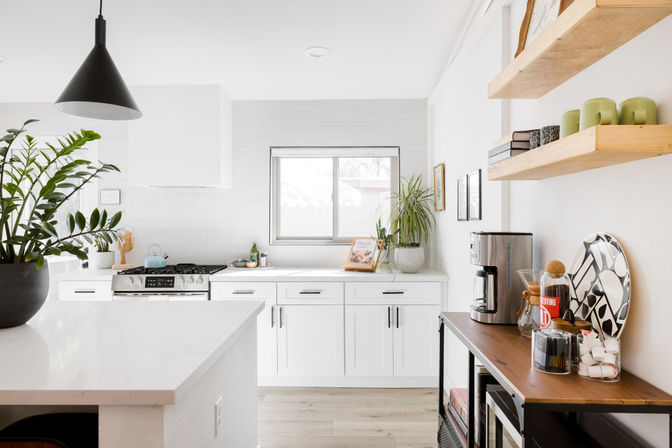 Bright sunlit modern white kitchen with a marble island, black pendant light, stainless steel range, potted plants by a window, open wood shelves, and a wooden coffee station with mugs and accessories.