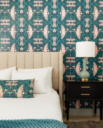 Whimsical bedroom scene with teal and pink bird-print wallpaper, beige upholstered headboard, white quilted bedding, matching teal bird-pattern lumbar pillow, chunky teal throw, black nightstand with brass pulls and green ceramic table lamp.