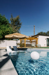 Sunny suburban backyard pool with sparkling blue water, a floating white ball, in-pool lounge chairs beneath a straw tiki umbrella, yellow wooden pergola and patio seating under a clear blue sky.