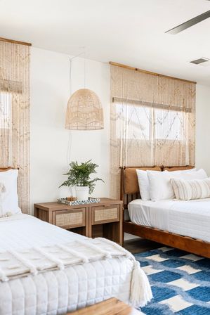Boho coastal bedroom with two wood-framed beds, white quilted bedding and textured pillows, rattan headboards and pendant light, macramé beaded curtains, wooden nightstand with potted plant, and a blue geometric rug.