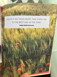 Close-up of a book page showing an inspirational quote, "Write it on your heart that every day is the best day in the year," above a sunlit wheat field sprinkled with red poppies, with a thumb holding the bottom edge.