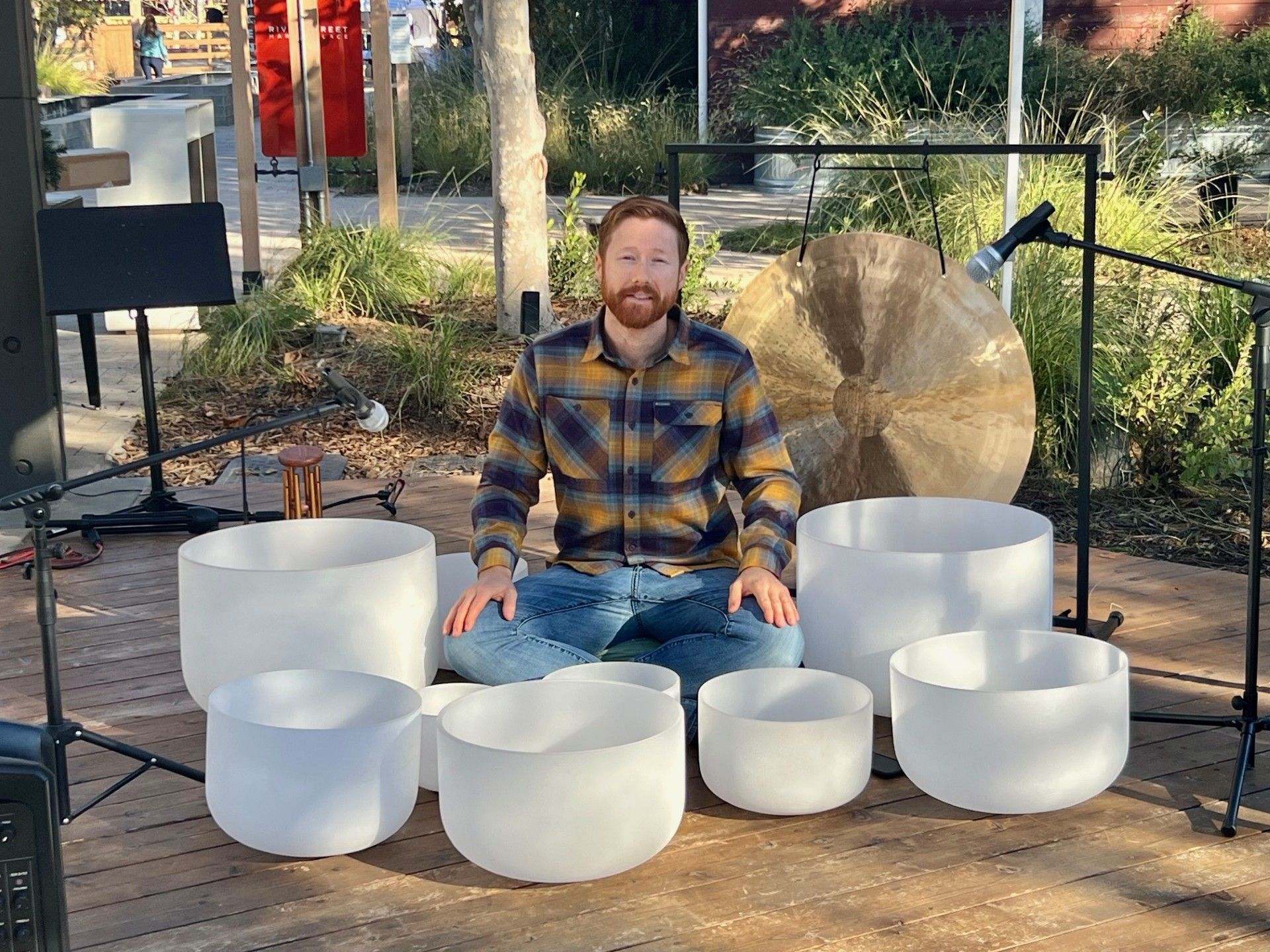 Person seated cross-legged on an outdoor wooden stage surrounded by white crystal singing bowls and a large bronze gong, with microphones and stands set up for a sound-healing performance.
