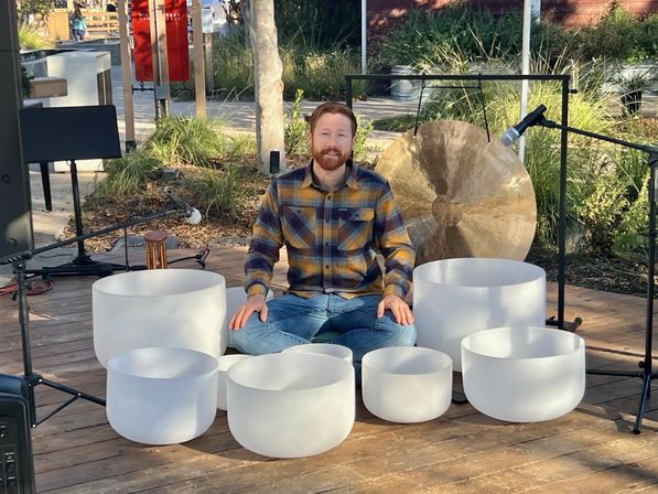 Person seated cross-legged on an outdoor wooden stage surrounded by white crystal singing bowls and a large bronze gong, with microphones and stands set up for a sound-healing performance.