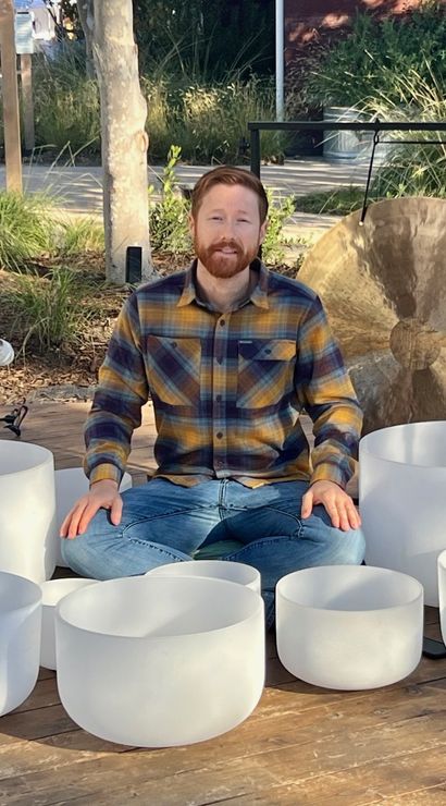Person seated cross-legged on an outdoor wooden stage surrounded by white crystal singing bowls and a large bronze gong, with microphones and stands set up for a sound-healing performance.