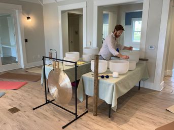 Person preparing a sound bath in a bright indoor wellness studio with crystal singing bowls on covered tables, mallets, a large gong on a stand, and yoga mats on a wood floor.