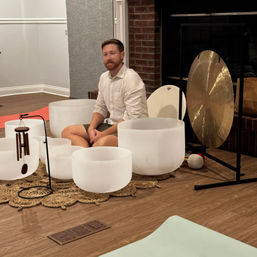 Person seated cross-legged among frosted crystal singing bowls, wind chimes and a large gong for a sound bath in an indoor meditation/wellness studio with wood floors.