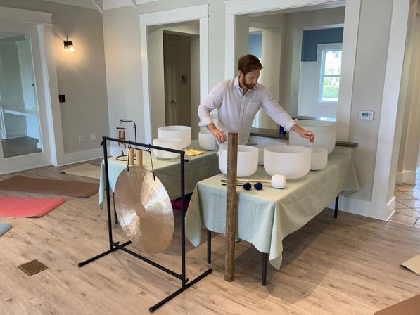 Person setting up crystal singing bowls and a gong for a relaxing sound bath in a bright wellness studio with yoga mats on a wood floor