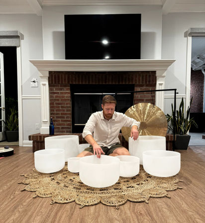 Person seated on a woven jute rug in a modern living room playing white crystal singing bowls with a large gong behind them, set in front of a brick fireplace and wall-mounted TV — intimate sound-bath meditation scene.