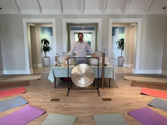 Bright indoor wellness studio with wooden floor, colorful yoga mats arranged in a circle and a person standing behind a large gong, singing bowls and mallets set up for a group sound bath/meditation session.