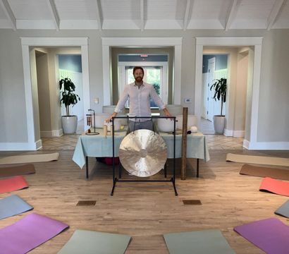 Bright indoor wellness studio with wooden floor, colorful yoga mats arranged in a circle and a person standing behind a large gong, singing bowls and mallets set up for a group sound bath/meditation session.