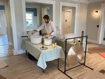 Person setting up crystal singing bowls and a large gong for a sound bath in a bright indoor wellness studio with yoga mats on a wood floor
