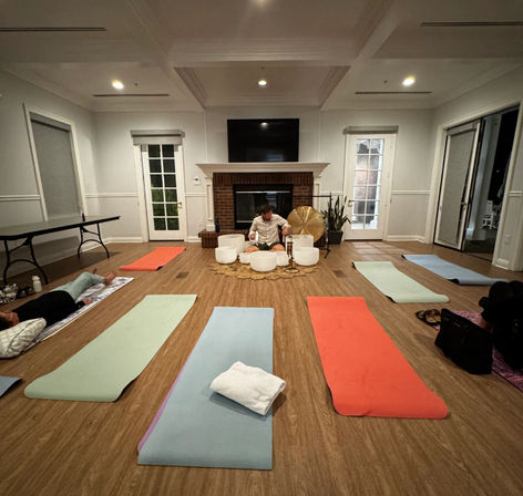 Cozy group sound-bath and yoga session with colorful mats on hardwood floor, participants relaxing while an instructor plays crystal singing bowls and a gong in front of a fireplace.