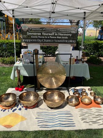 Outdoor park sound-healing booth under a white canopy showing a large gong, rows of ornate metal singing bowls, crystal bowls, mallets and cushions arranged on a patterned rug — wellness, meditation and sound therapy display at a sunny community market.