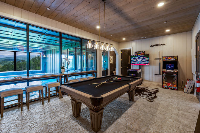 Game room with black-felt pool table, hanging globe pendant lights, arcade machines and wall-mounted dartboard, bar seating overlooking an indoor pool and mountain view through large glass windows, wood-paneled ceiling and textured stone floor.