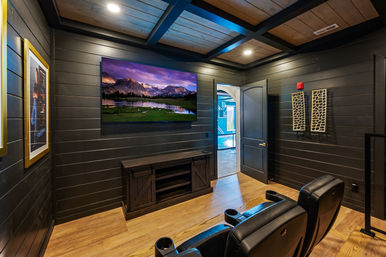 Cozy home theater with black shiplap walls and wood-beamed ceiling, wall-mounted TV showing a mountain scene, two black leather recliners, rustic media console, and an open door leading to an indoor pool area.