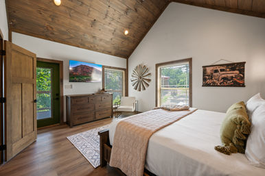 Sunlit rustic cabin bedroom with vaulted wood ceiling, king bed with green throw pillow and beige quilt, wooden dresser with wall TV, decorative windmill, windows and open door to leafy porch.