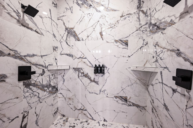 Spa-like white marble shower with bold gray veining, dual black rain showerheads and controls, corner shelves and three wall-mounted soap dispensers