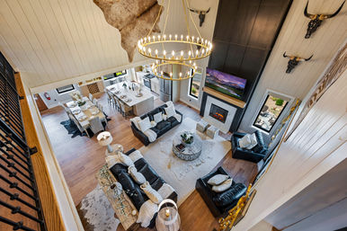 Aerial view of a two-story modern farmhouse open-concept living room and kitchen with vaulted shiplap ceiling, large circular chandelier, stone fireplace and black leather sofas.