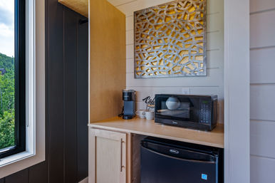Cozy cabin kitchenette with a wood countertop holding a black microwave, mini fridge below, single‑serve coffee maker and mugs, geometric metal wall art above, and a window with lush green hillside view.