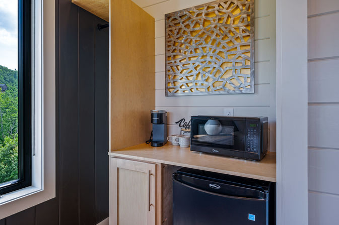 Cozy cabin kitchenette with a wood countertop holding a black microwave, mini fridge below, single‑serve coffee maker and mugs, geometric metal wall art above, and a window with lush green hillside view.