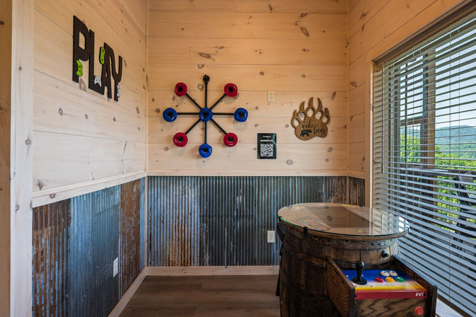 Rustic cabin game corner with light pine walls and corrugated metal wainscoting, colorful wall ring-toss game and 'PLAY' letters, bear-paw decor, barrel-style arcade game with glass top, and blinds revealing a green mountain view.