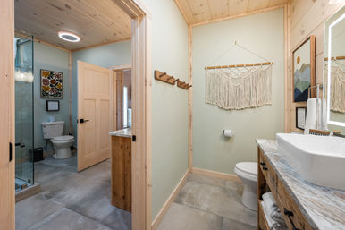 Cozy modern rustic bathroom with light wood trim and ceiling, marble-top vanity with vessel sink, glass-enclosed shower, toilet in adjoining room, macramé wall hanging, wooden towel hooks and large gray tile floor.