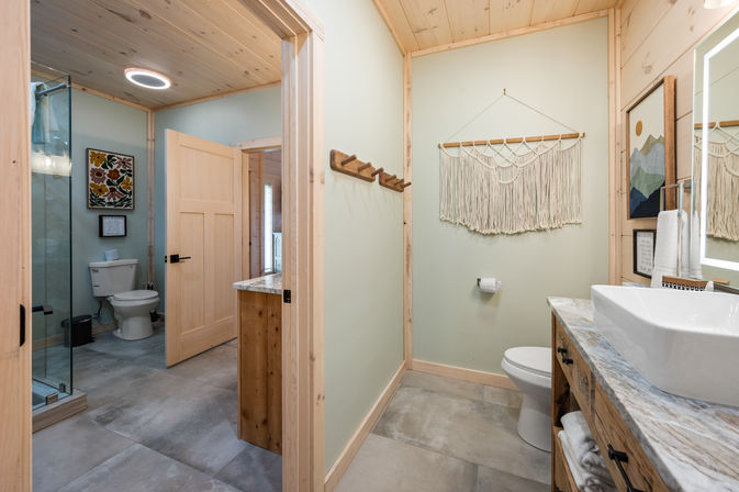 Cozy modern rustic bathroom with light wood trim and ceiling, marble-top vanity with vessel sink, glass-enclosed shower, toilet in adjoining room, macramé wall hanging, wooden towel hooks and large gray tile floor.