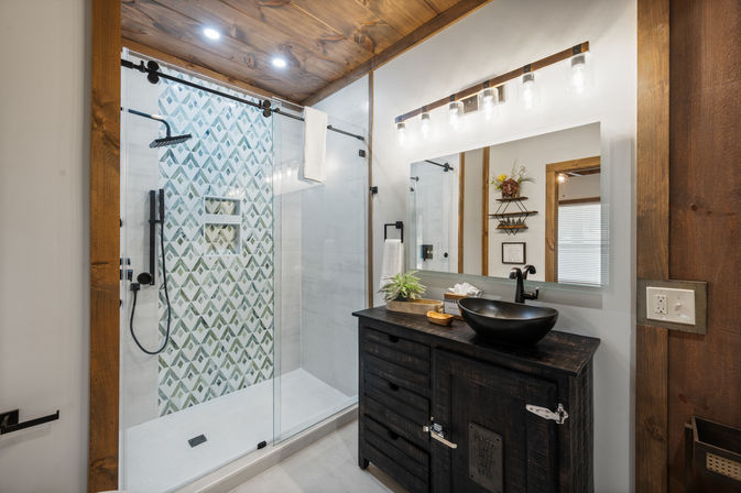 Cozy modern-rustic bathroom interior with glass walk-in shower featuring green geometric accent tile and matte-black rain shower, wood-paneled ceiling, dark reclaimed-wood vanity with black vessel sink, and illuminated mirror.