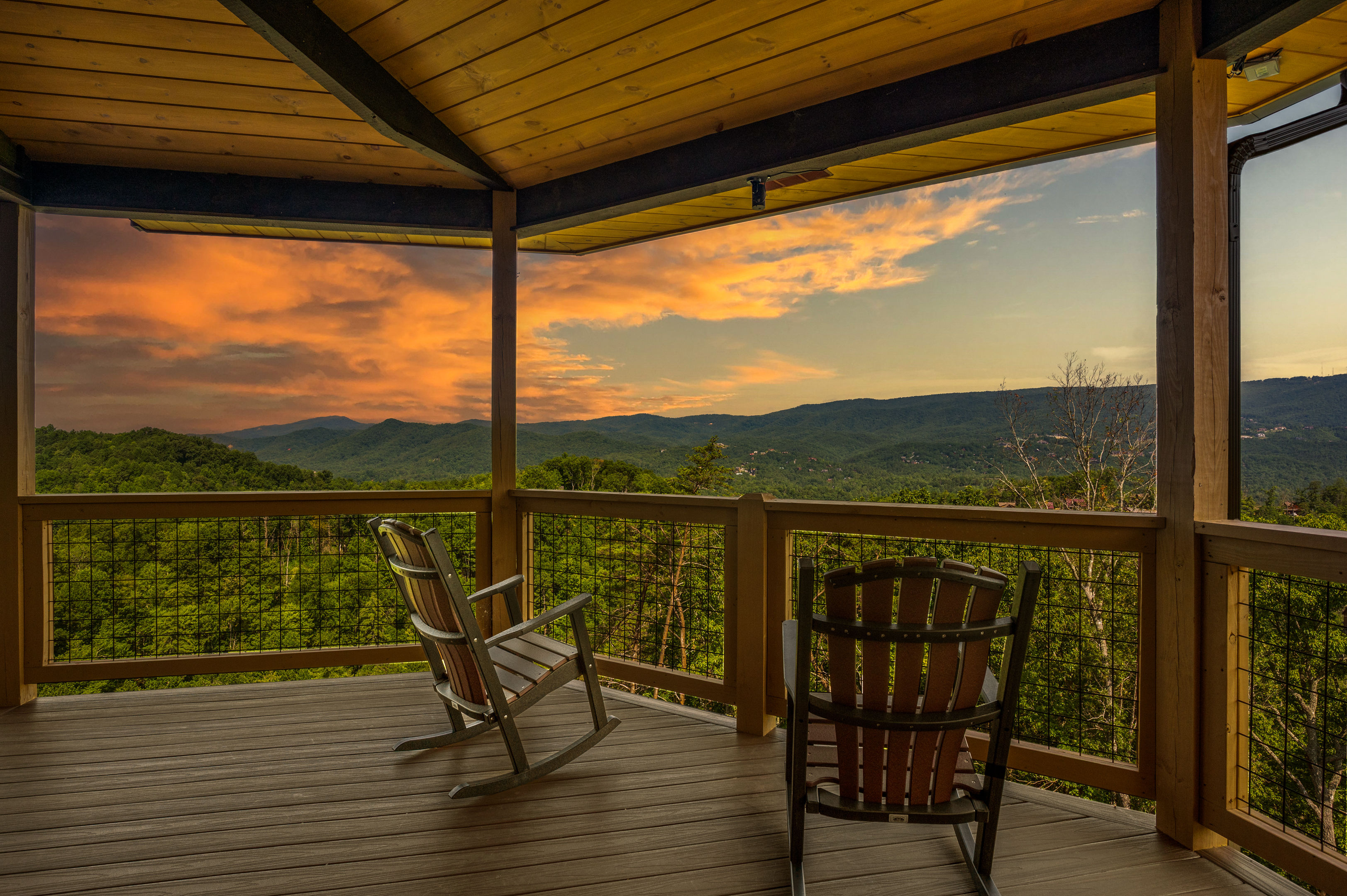 Cozy covered wooden deck with two rocking chairs overlooking green, forested mountains and a golden-orange sunset sky — panoramic mountain view.